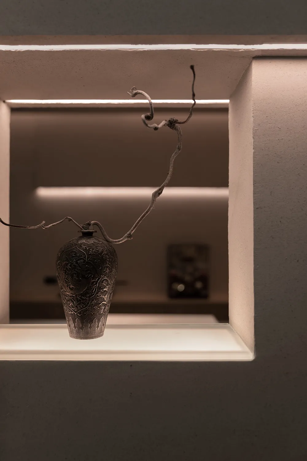 A dimly lit room with a metal vase on a table against a white wall, minimalist composition