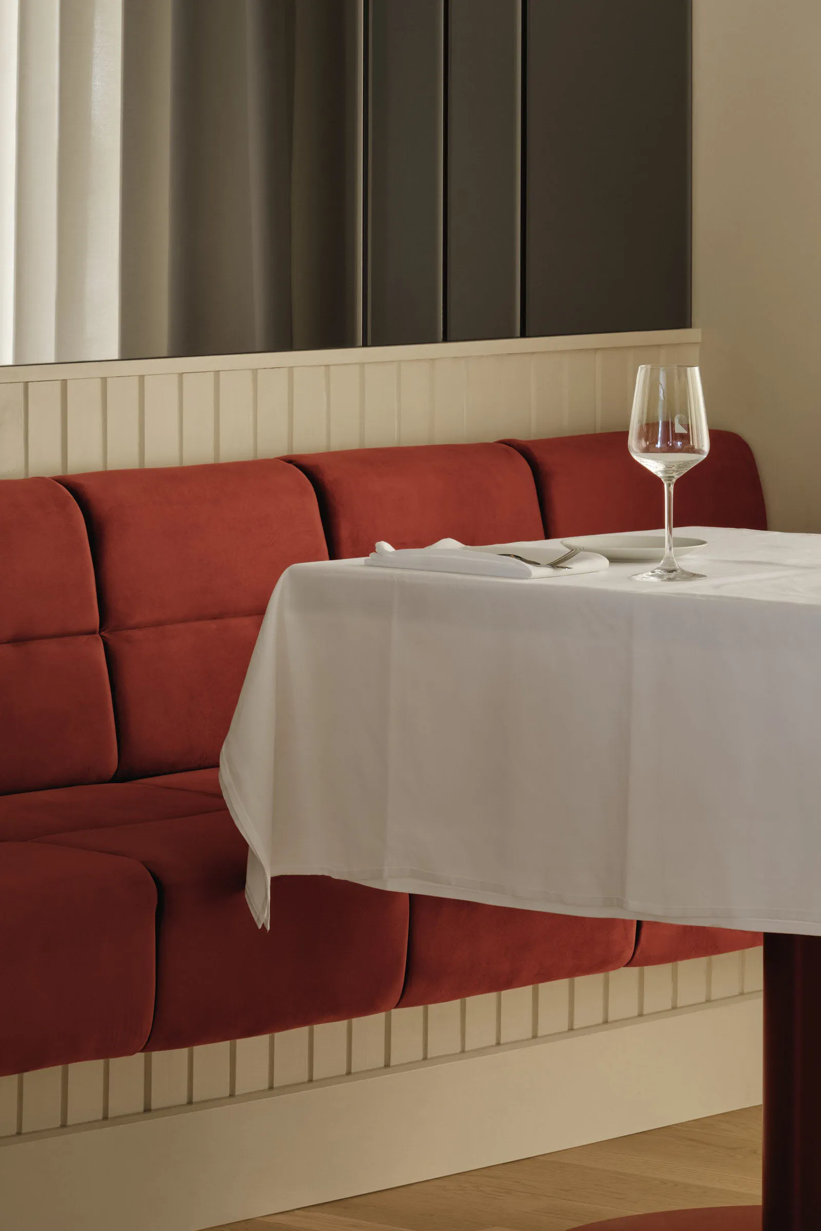 A dining table with a white tablecloth and wine glass near a window, surrounded by a red and black couches in a restaurant setting
