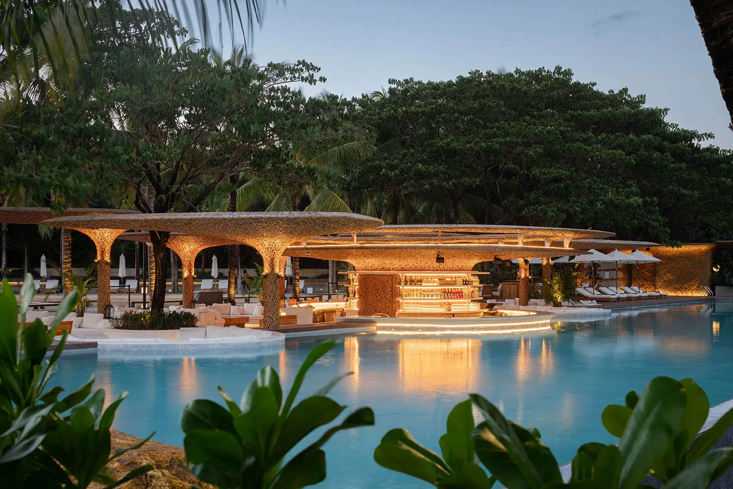 A resort pool area at night surrounded by palm trees, with chairs and umbrellas for relaxation and socializing