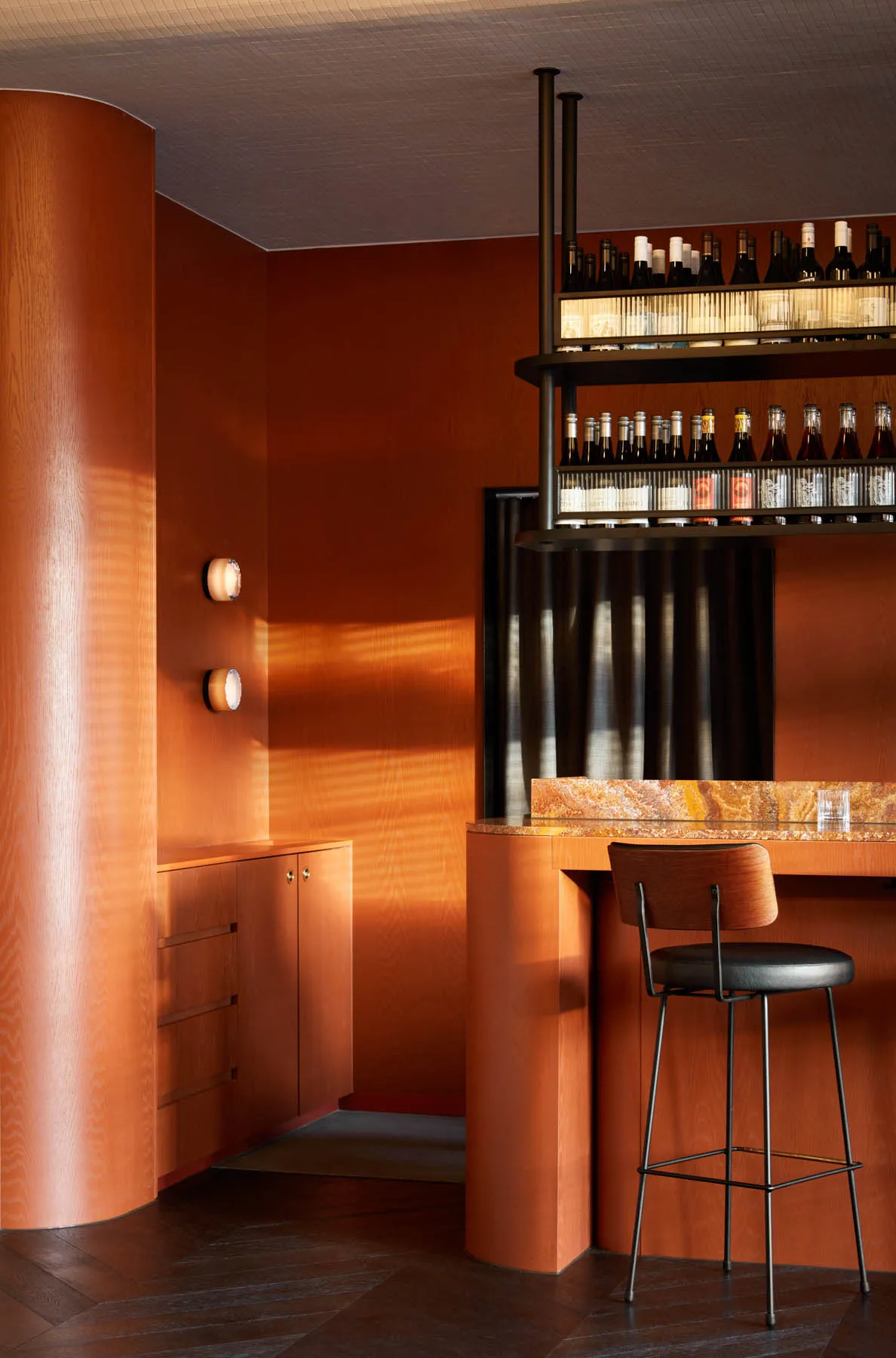 A cozy bar area with a red-painted wall, a counter, a stool, and a wine rack with numerous bottles, creating a warm and inviting atmosphere for patrons