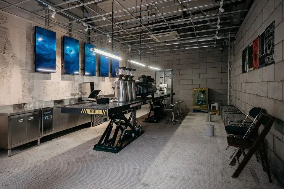 Industrial kitchen with concrete floors and walls, featuring a countertop with various items and chairs, lit by overhead lighting