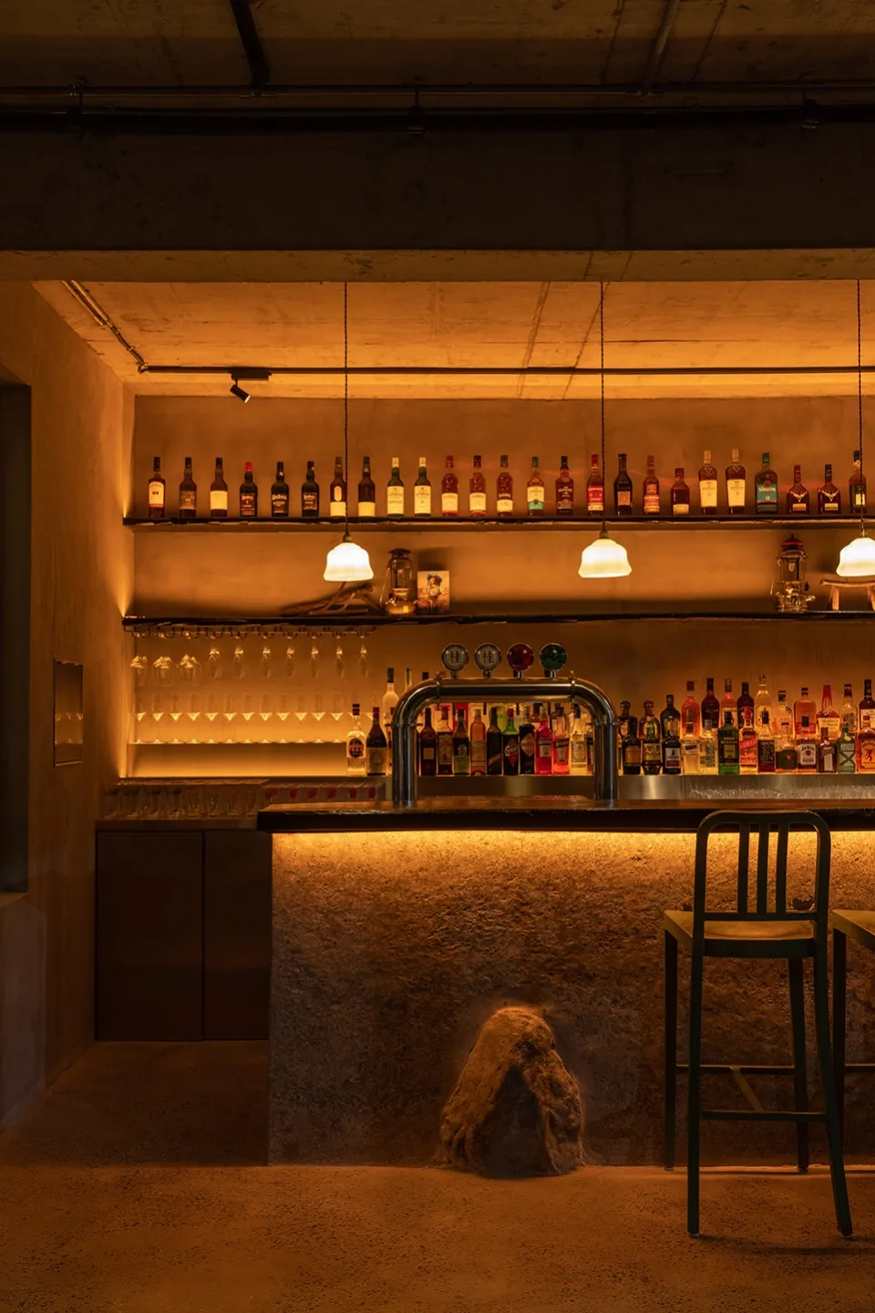A cozy bar area with a long wooden counter featuring bottles and wine glasses, accompanied by two chairs in a warm and inviting atmosphere