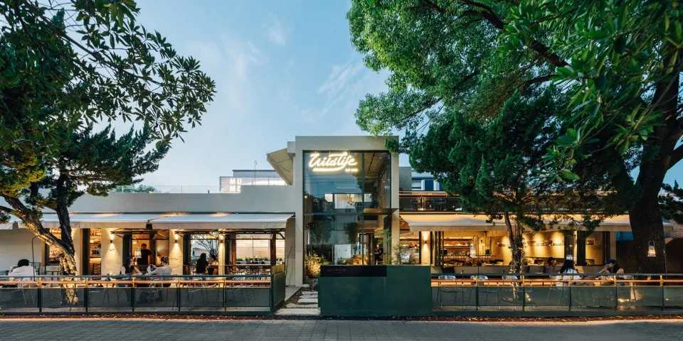 A modern restaurant with a green building, surrounded by trees, featuring dining tables, chairs, and potted plants, conveying a warm and welcoming ambiance