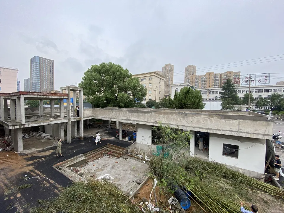 A busy construction site with a large concrete building under construction, featuring multiple floors, a rooftop area, and workers scattered throughout