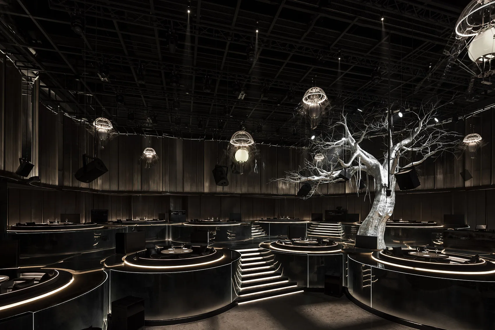 A modern event space with a tree sculpture on the stage, surrounded by tables and chairs, and multiple dining tables with chairs nearby