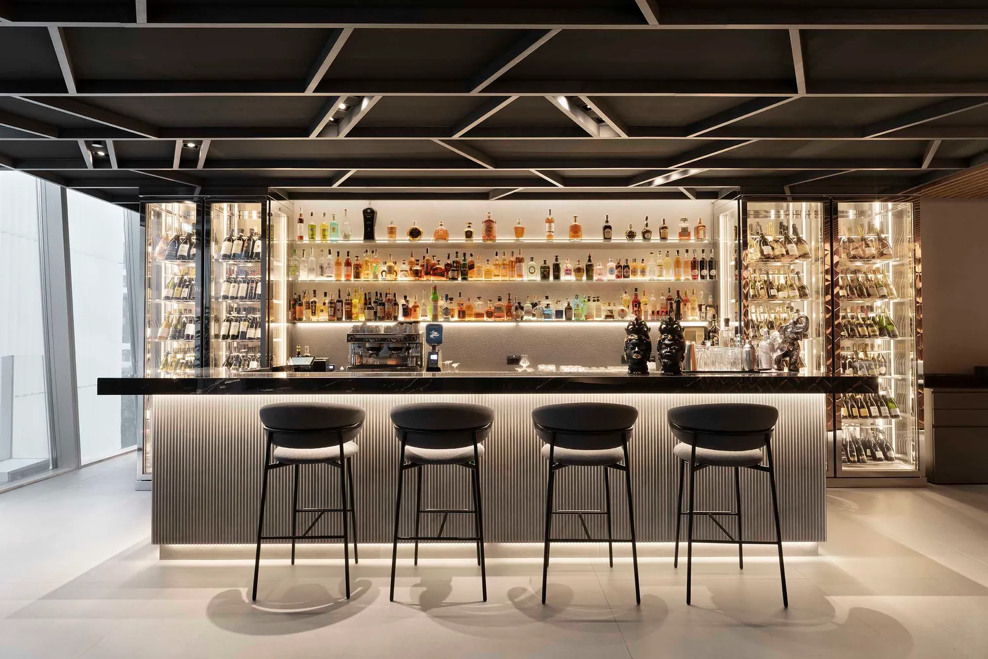 A modern bar with a long counter and four stools, featuring numerous bottles and a stylish design