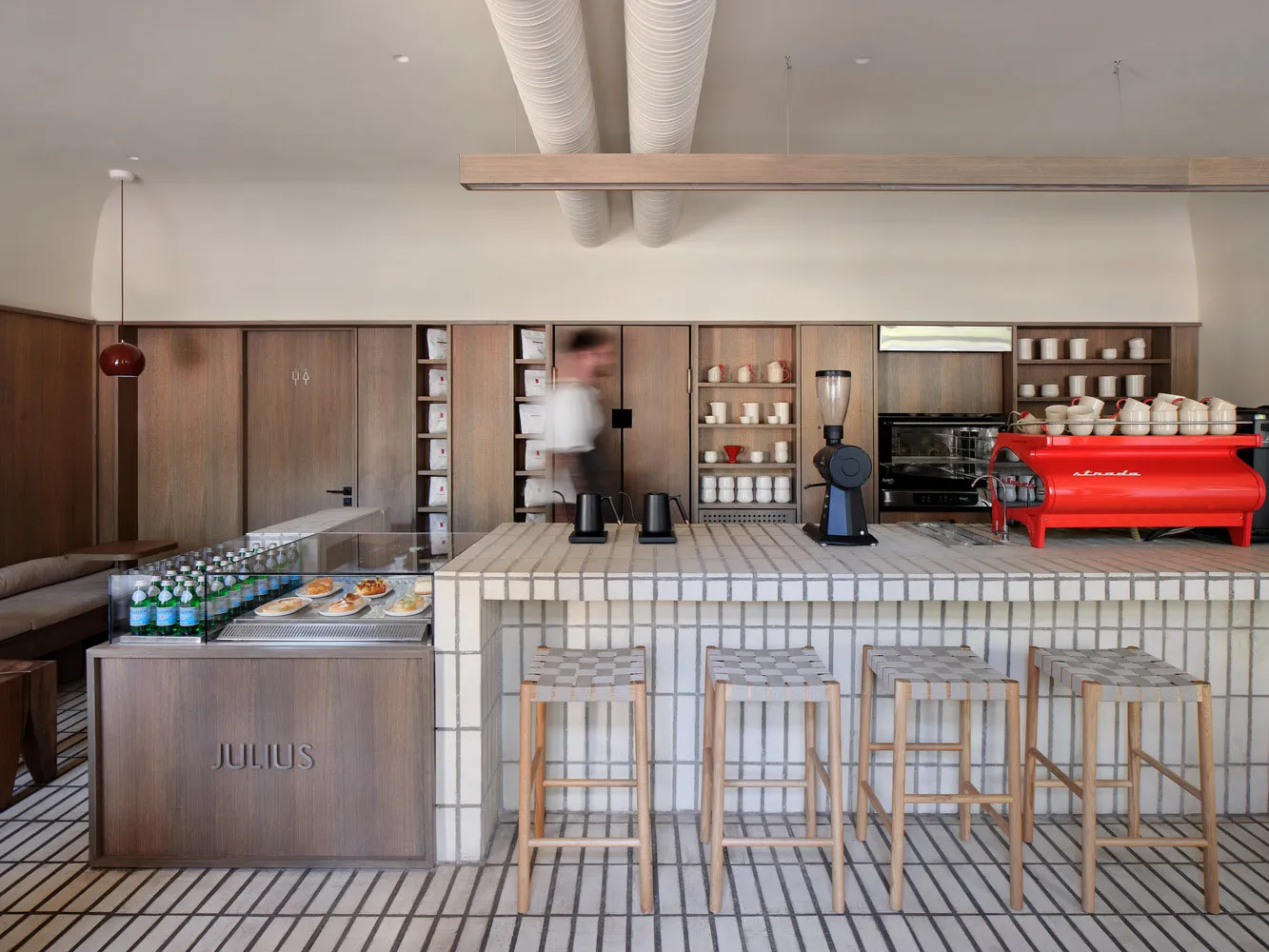 Casual restaurant kitchen with wooden cabinets and counters, two dining tables, and various cups and bottles scattered throughout, with a person in the background