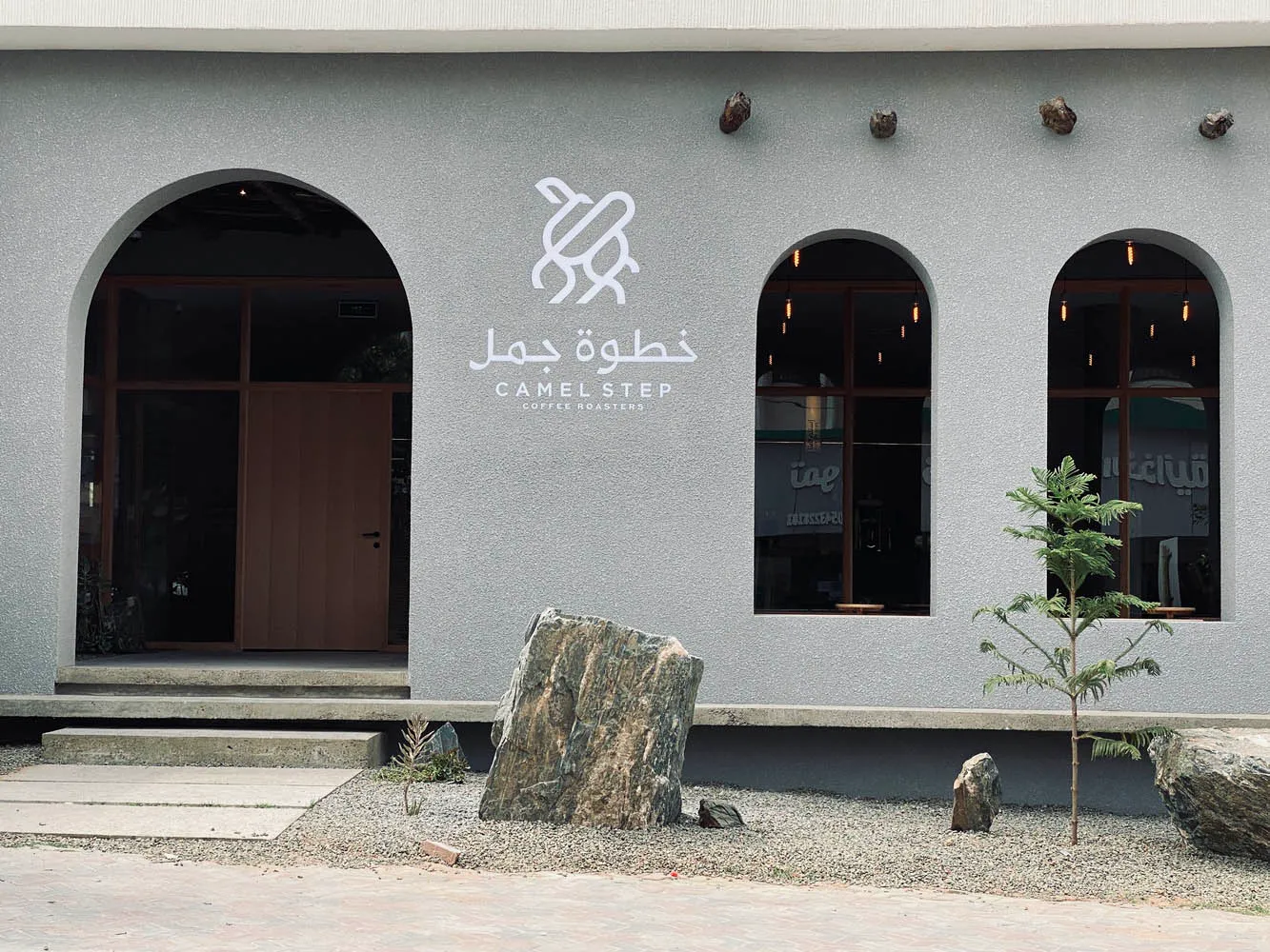 A modern building with a gray color scheme and a stone wall in front of a doorway, surrounded by two trees and a serene atmosphere