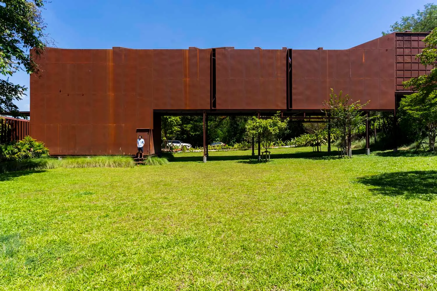 A relaxed outdoor space with a metal structure in the background, benches scattered throughout, and lush greenery adding to the ambiance