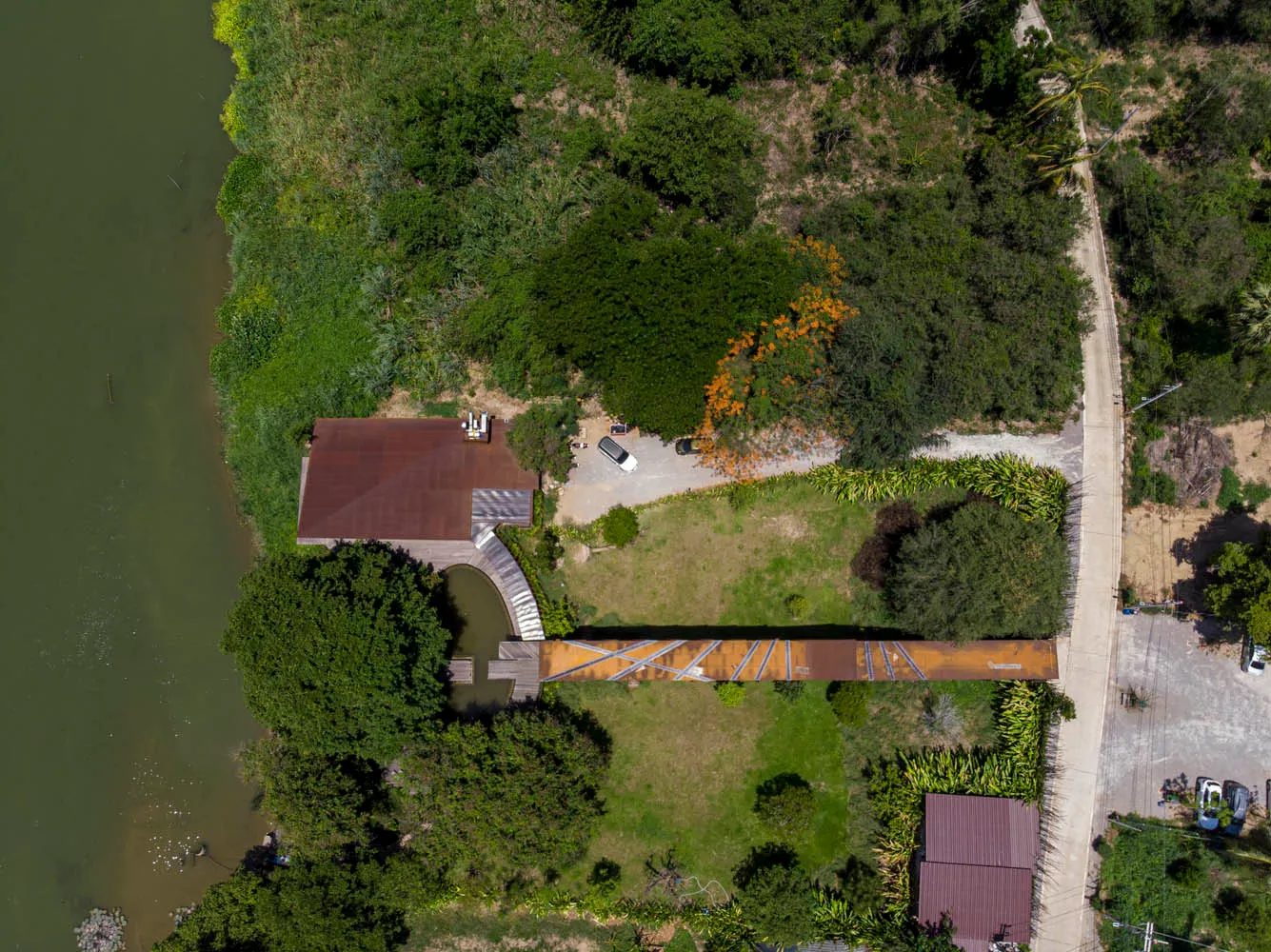 A house with a brown roof and a wooden bridge overlooking a river, surrounded by trees and a parked car