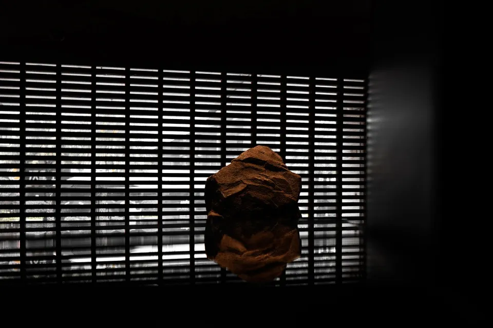 A brown rock sits on a reflective glass table in an indoor space with vertical blinds on the wall