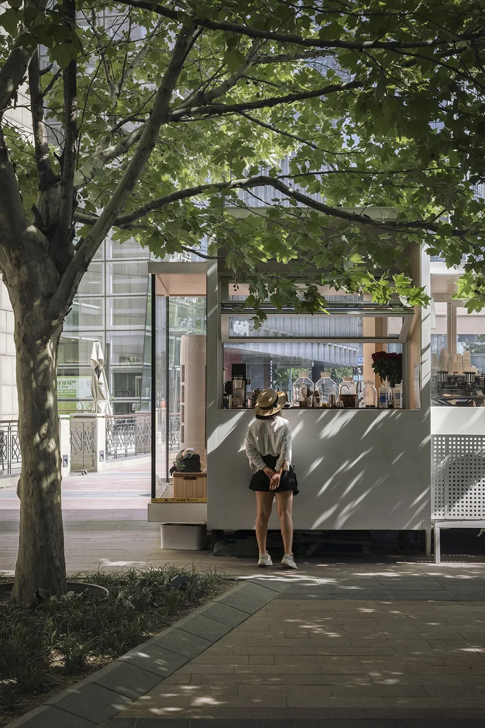 A casual outdoor cafe scene with a woman standing in front of a small white food stand, surrounded by potted plants and other people in the background