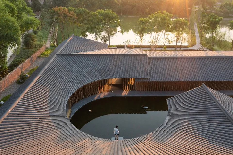 寧靜的建築景觀，圓形結構周圍環繞樹木與水景，結構內平台上站著一人