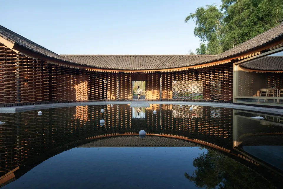 A serene Asian-style building with a wooden roof surrounded by trees, featuring a courtyard with a pond and several people in a peaceful environment