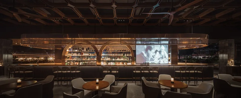 A cozy bar area with a wooden counter, chairs, and a mounted TV, surrounded by bottles and inviting atmosphere