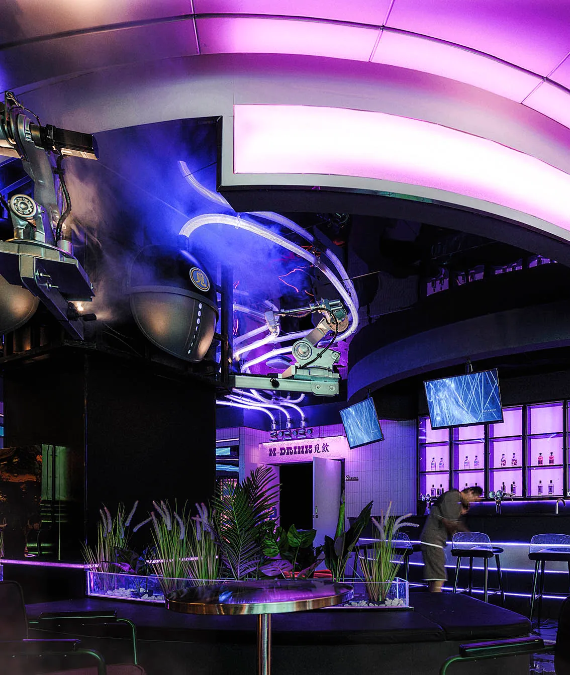 A modern bar area with a purple ambiance, featuring a circular table, chairs, potted plants, and a sleek bar with stainless steel or contemporary materials