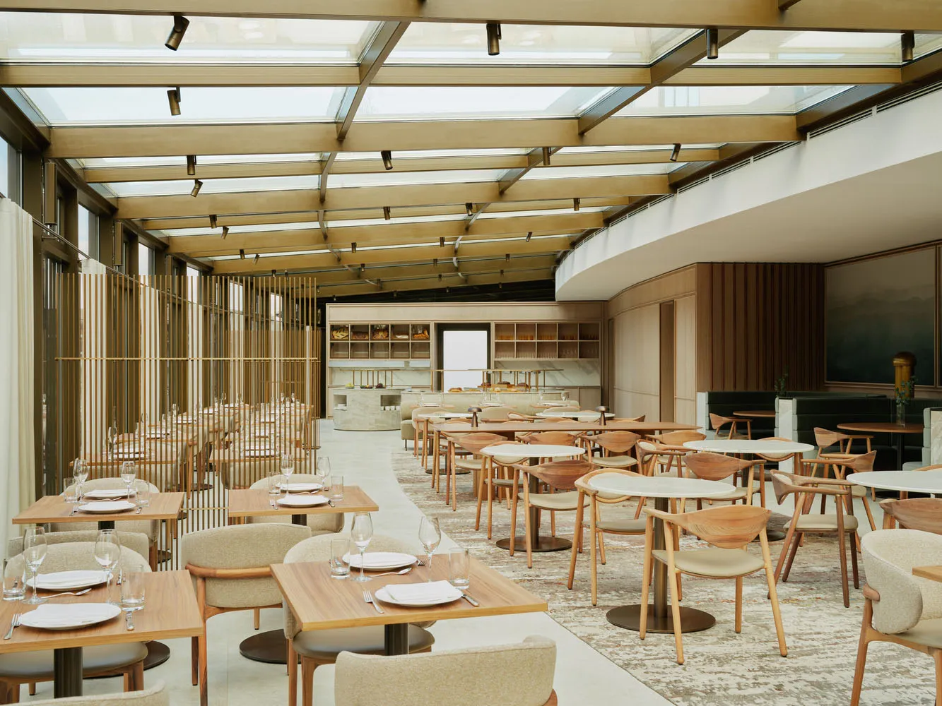 A spacious restaurant interior with wooden tables and chairs, natural light from an open ceiling, and wine glasses on some tables
