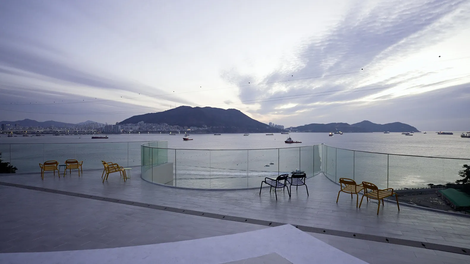 A patio area with chairs and dining tables arranged around a glass wall overlooking the ocean, creating a relaxing and inviting atmosphere