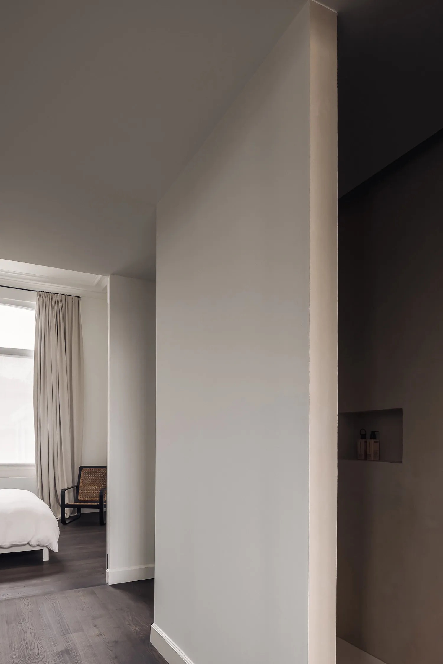 A dimly lit bedroom with a white bed and two chairs, surrounded by white walls and a minimalist aesthetic