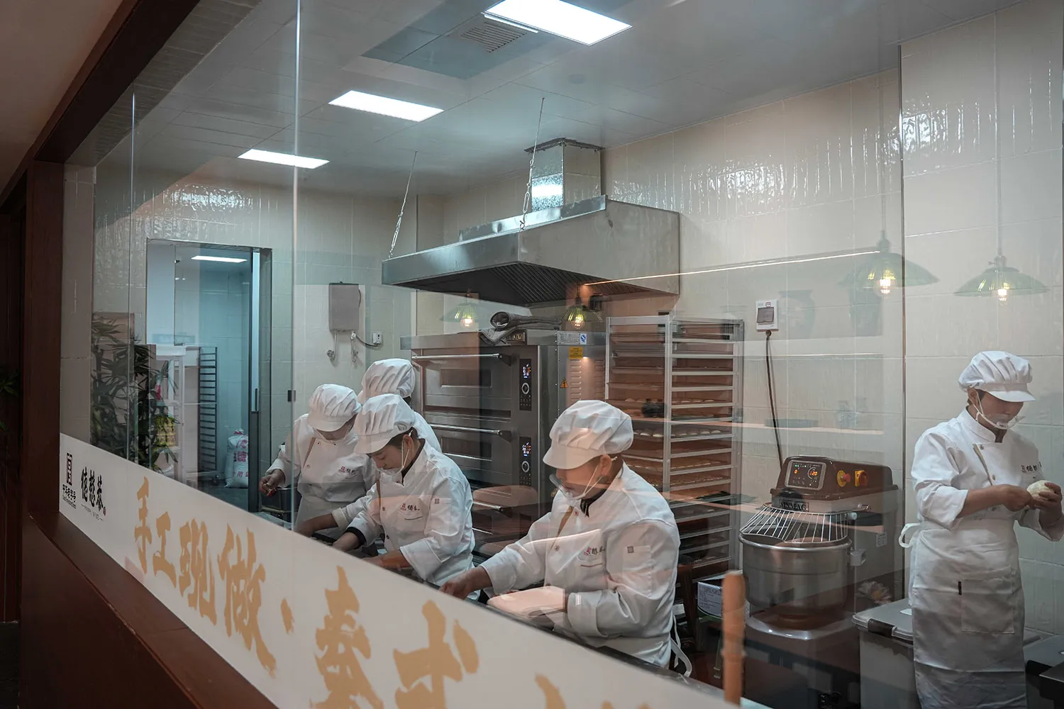 專業廚房場景，四名戴白帽工人隔著玻璃屏風準備食物，周圍環繞廚房家電