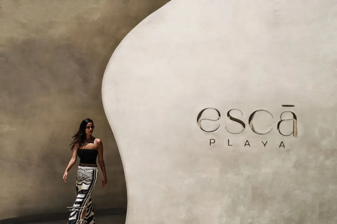A woman in a black dress walks past a wall with an advertisement for Escapade Playa in a modern and stylish hotel lobby