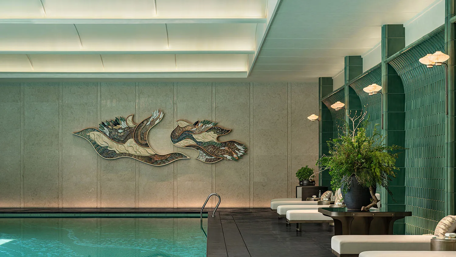 A large indoor pool area with a mosaic tile design on the walls, surrounded by seating and potted plants, creating a tranquil atmosphere