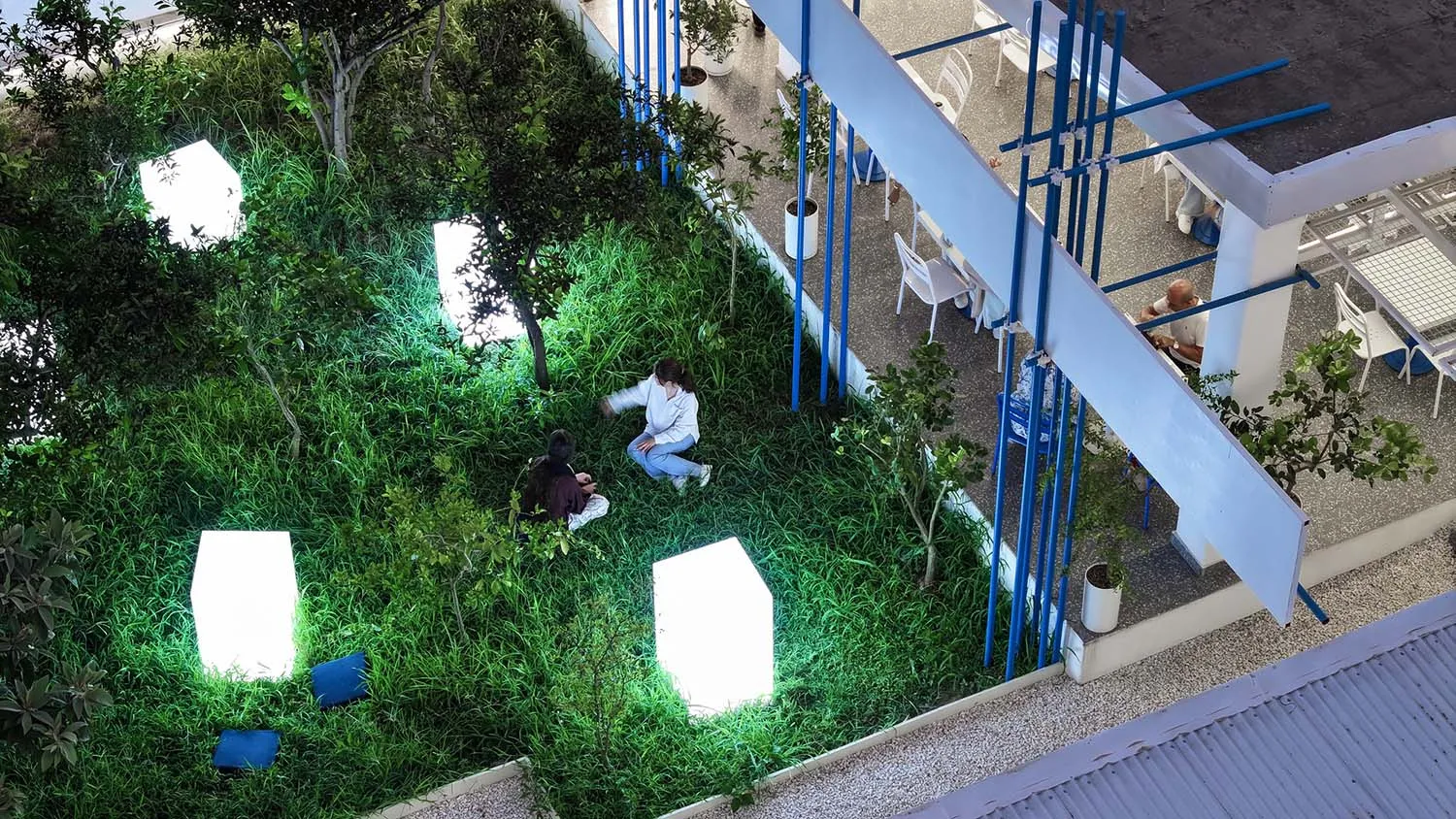 A serene indoor garden with grass, trees, and plants, featuring two people sitting on the grass, surrounded by lush greenery and artificial lighting