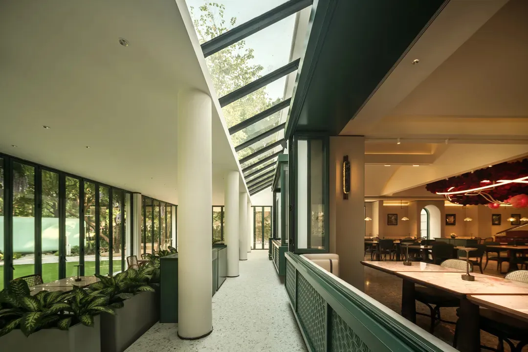Modern restaurant interior with green walls, glass ceiling, and natural light, featuring dining tables and chairs, and potted plants