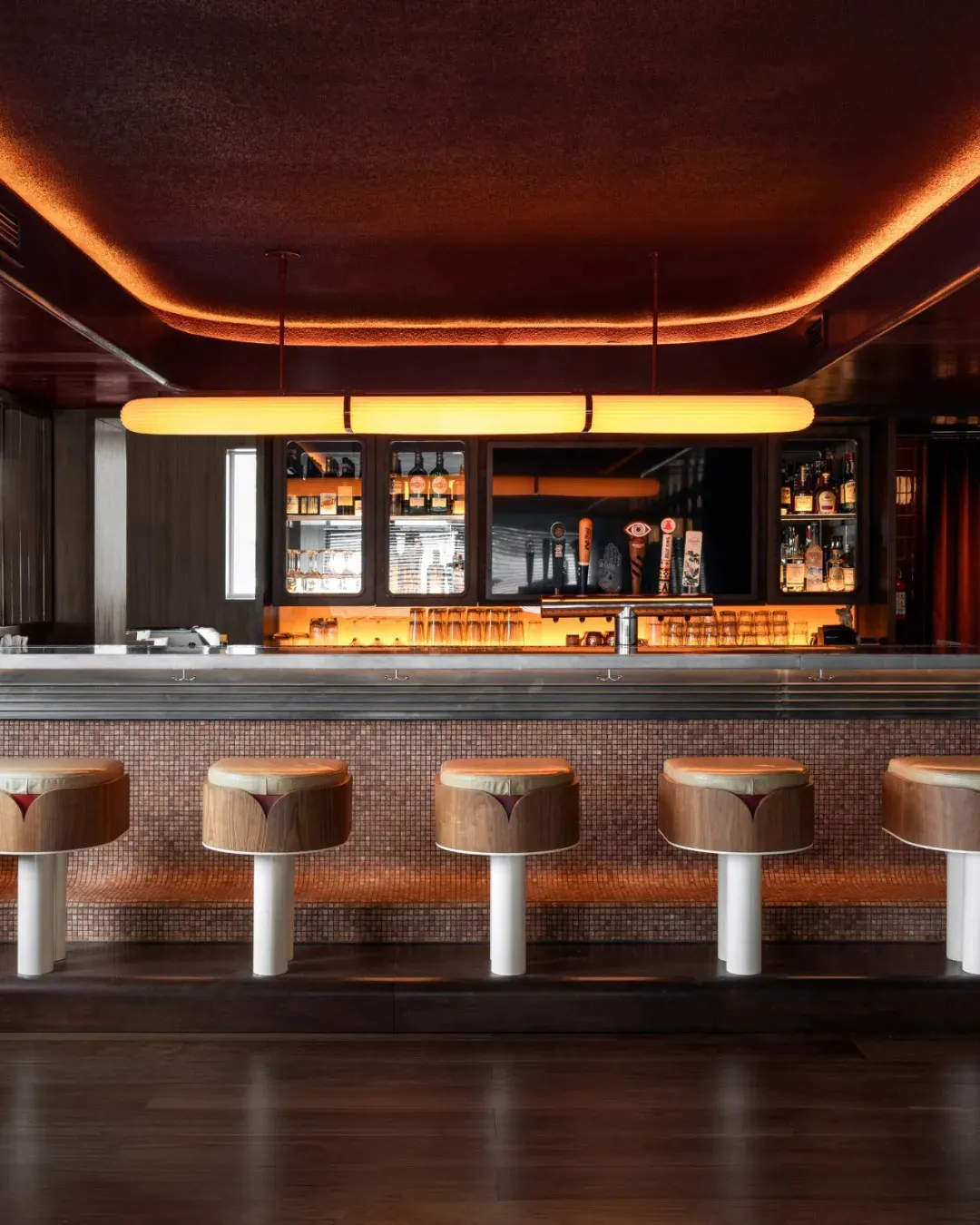 A bar area in a restaurant with a long wooden counter, stools, bottles, and cups, creating an inviting atmosphere for patrons to relax and socialize