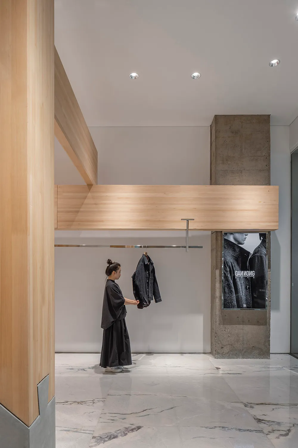 A woman holds up a black jacket in a well-stocked retail store with a concrete wall and various clothing items on display