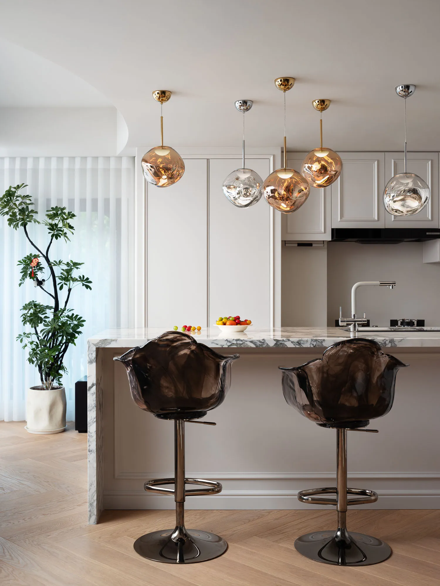 A modern kitchen with white walls, marble countertops, and stainless steel appliances, featuring a cozy dining area with barstools and a pendant light fixture, and a display of fresh fruits on the countertop