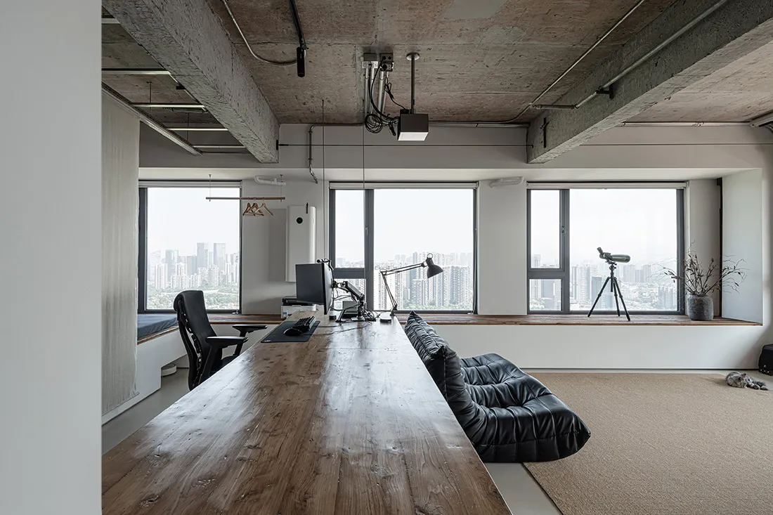 Modern living space with a large window overlooking a cityscape, featuring a wooden table, couch, chair, and two TVs in a bright and inviting atmosphere