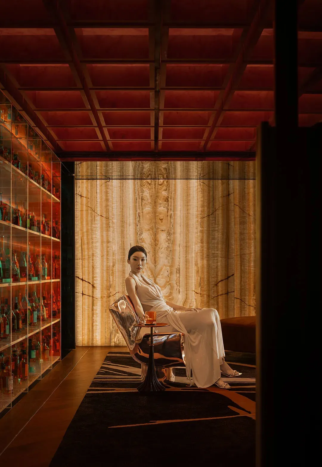 A woman sits in a chair in a well-decorated living area with bottles and wine glasses, conveying a sense of comfort and refinement