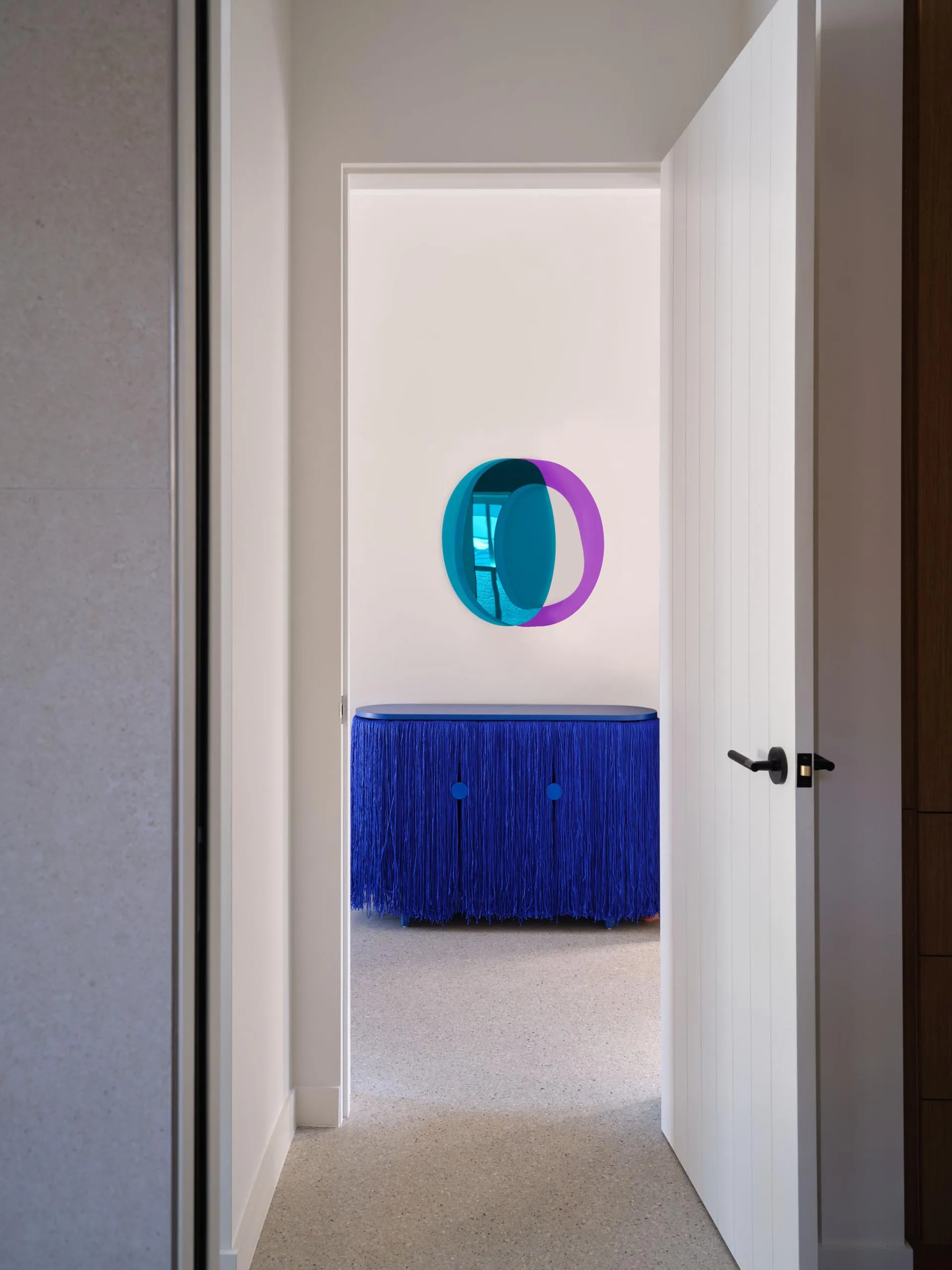 Cozy living room with a blue couch in front of a mirror with a purple circle design, two chairs, and a doorway leading to another room