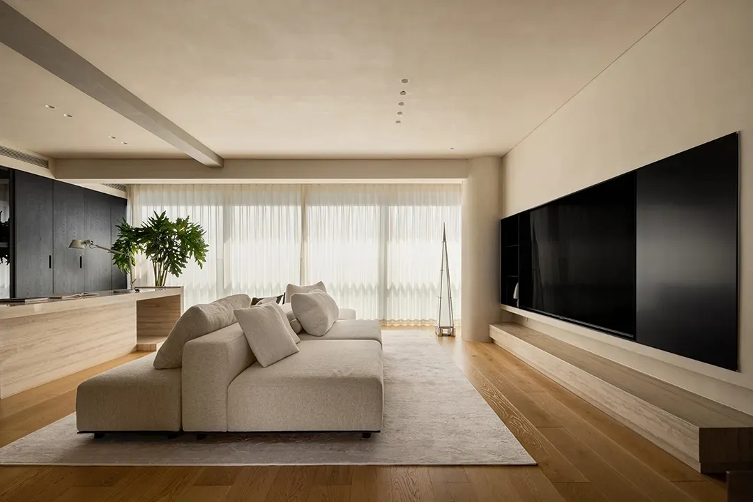 A spacious living room with a large couch, two chairs, a mounted television, and a potted plant, set against a backdrop of white walls and natural light