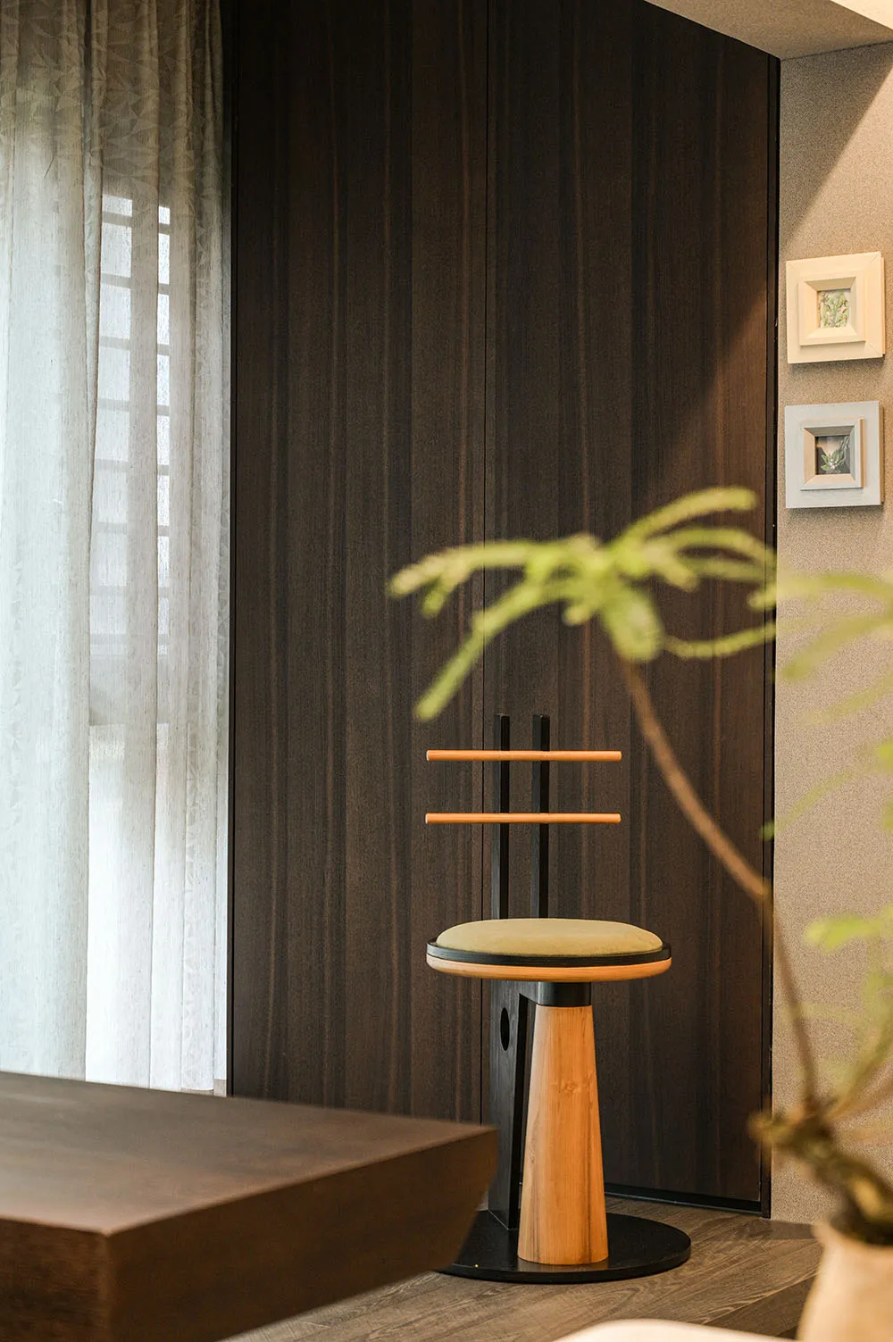 A wooden chair sits in front of a window with two potted plants on either side, surrounded by three framed pictures on the wall