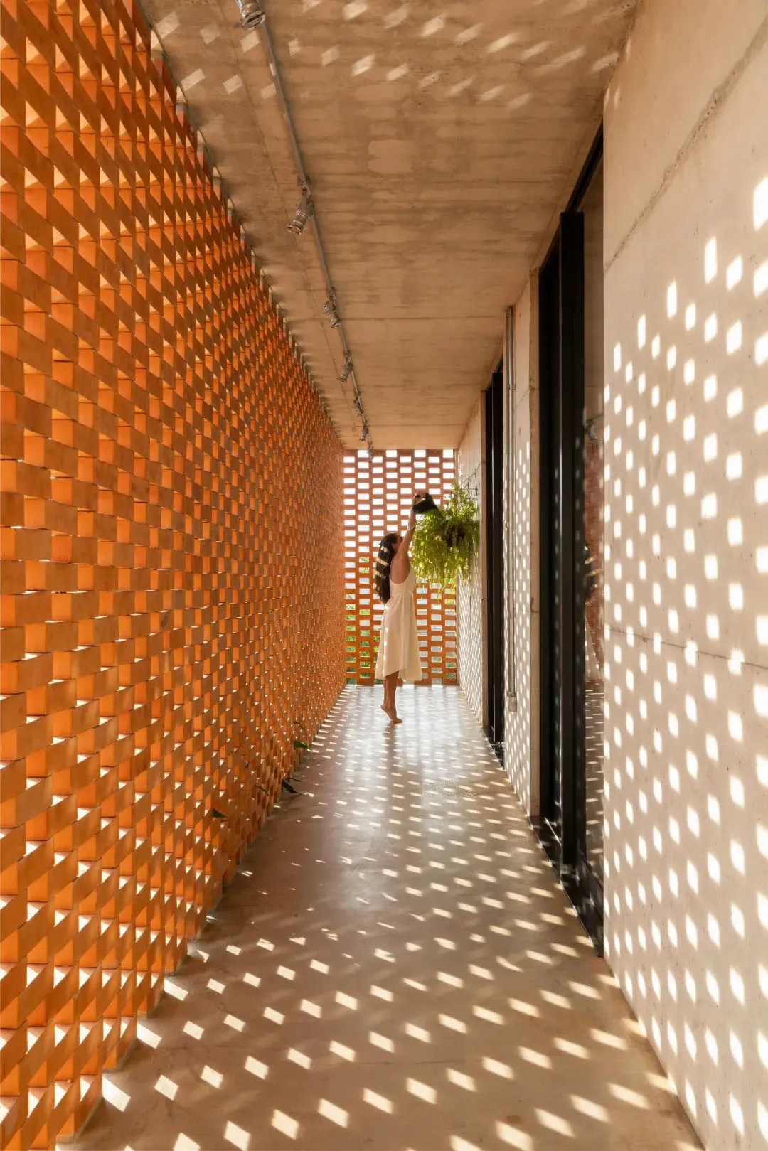 A woman stands in a white tile hallway with a potted plant on the wall, looking at it while holding a pole, with sunlight illuminating the space through windows and two sets of doors visible