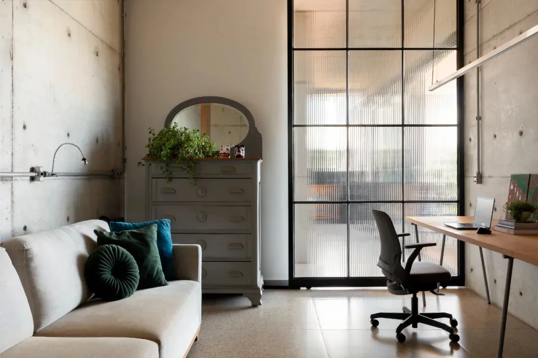 A minimalist living room with a couch, chair, desk, and dresser, surrounded by white walls and a large mirror, with wood and metal furniture