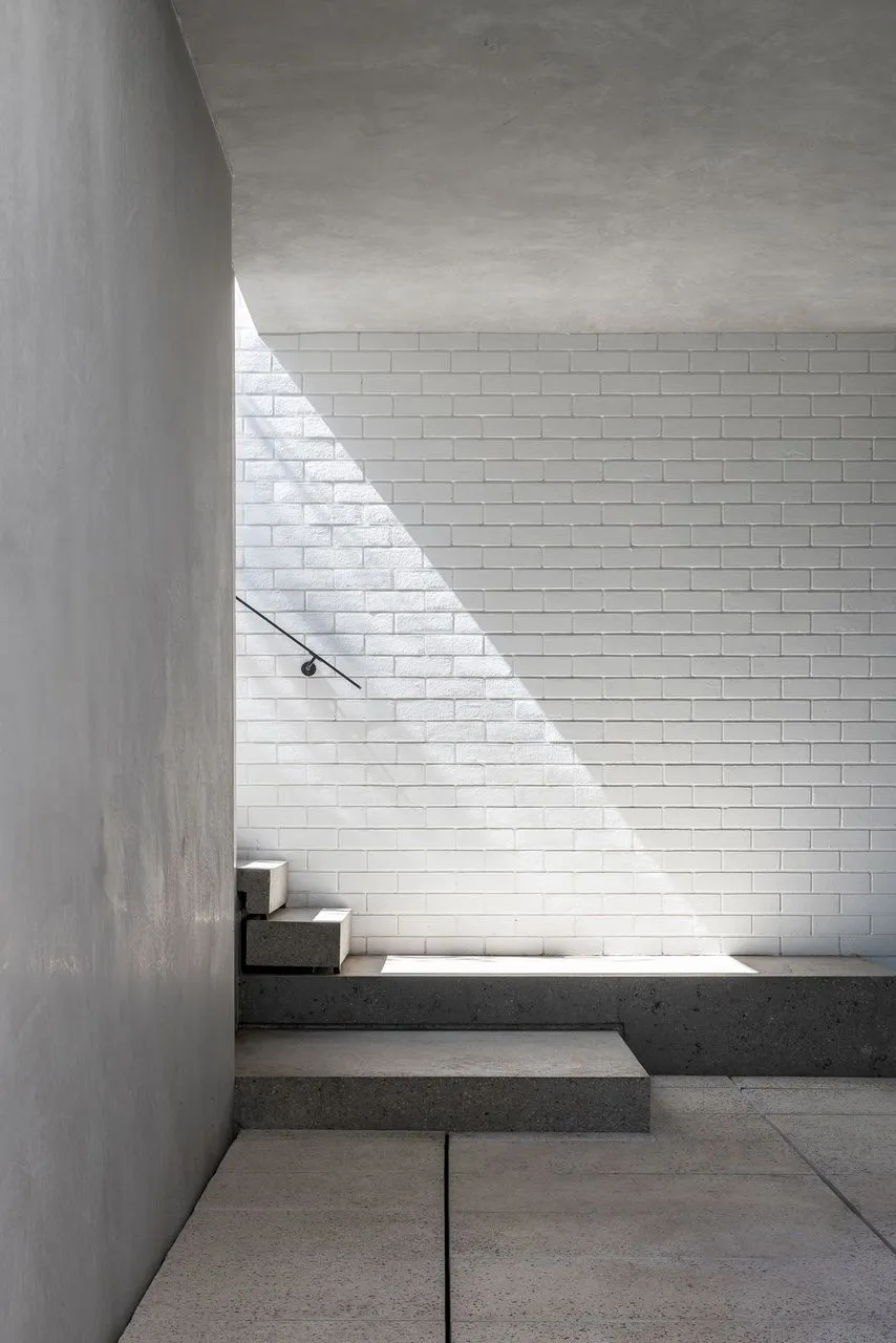 A minimalist staircase with a handrail leads to a white brick wall, set against a possibly residential or commercial building exterior