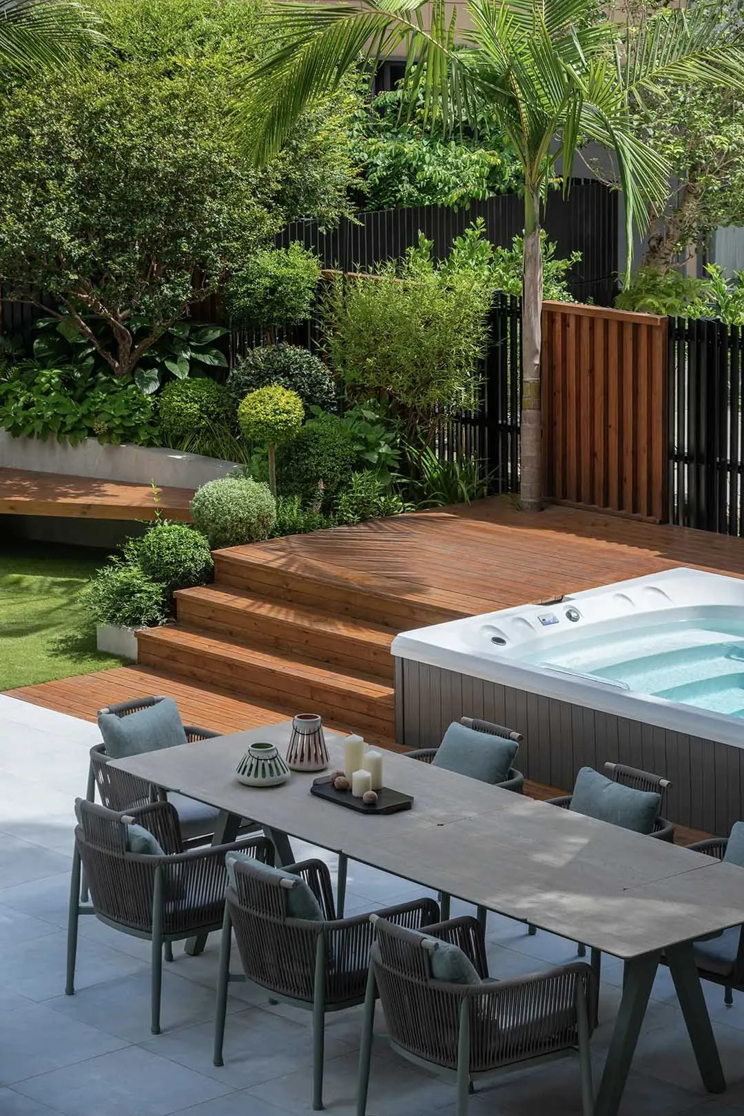 A relaxing outdoor patio area with a wooden deck, hot tub, and dining table surrounded by chairs, surrounded by lush greenery and potted plants