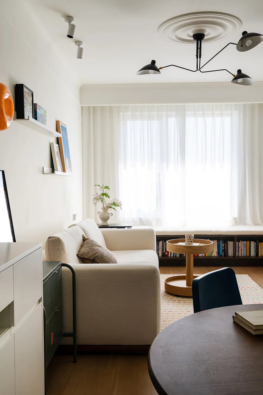 Bright and inviting living room with white walls, a couch, chair, dining table, and bookshelf, with sunlight streaming through the windows