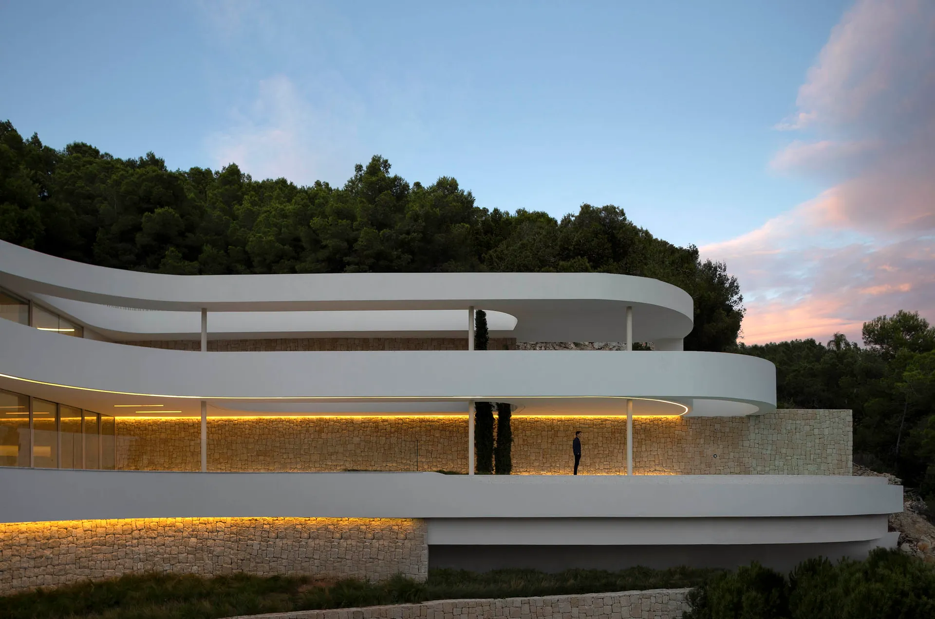 A modern building with a curved design and white exterior, featuring a balcony and surrounding trees