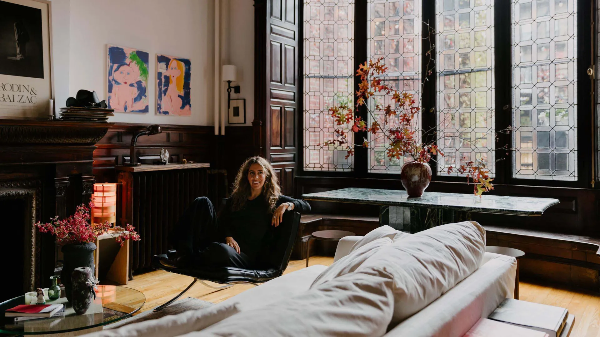 A well-decorated living room with a woman sitting on a chair in front of a window, surrounded by couches, a dining table, potted plants, and natural light
