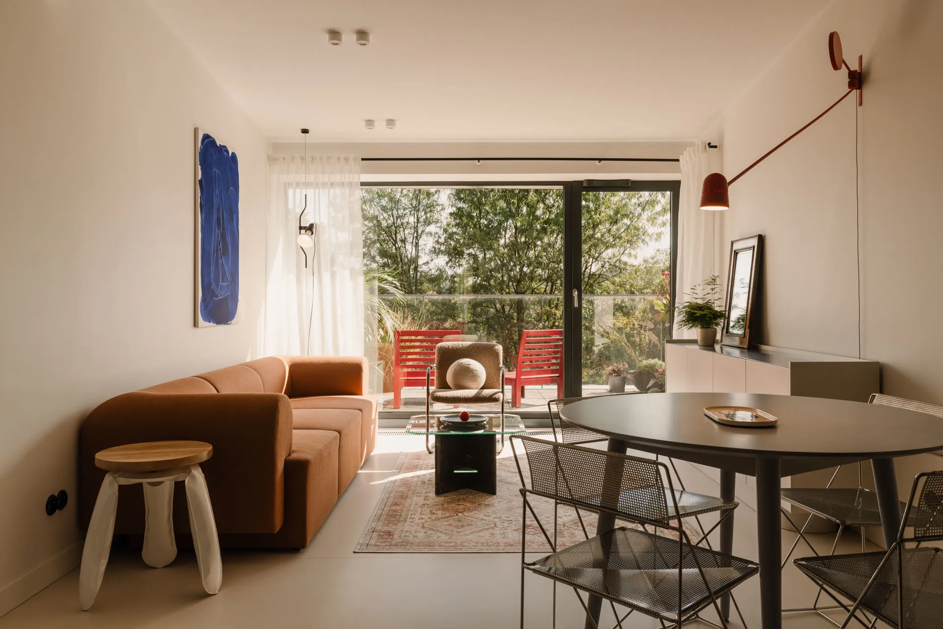 Open living area with couches, chairs, and a dining table, surrounded by greenery and brightly lit