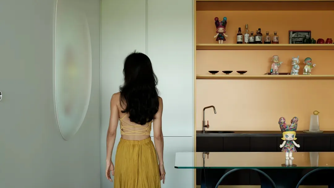 A woman stands in a kitchen with a sink, dining table, and bottles, surrounded by two chairs in a cozy and functional living space