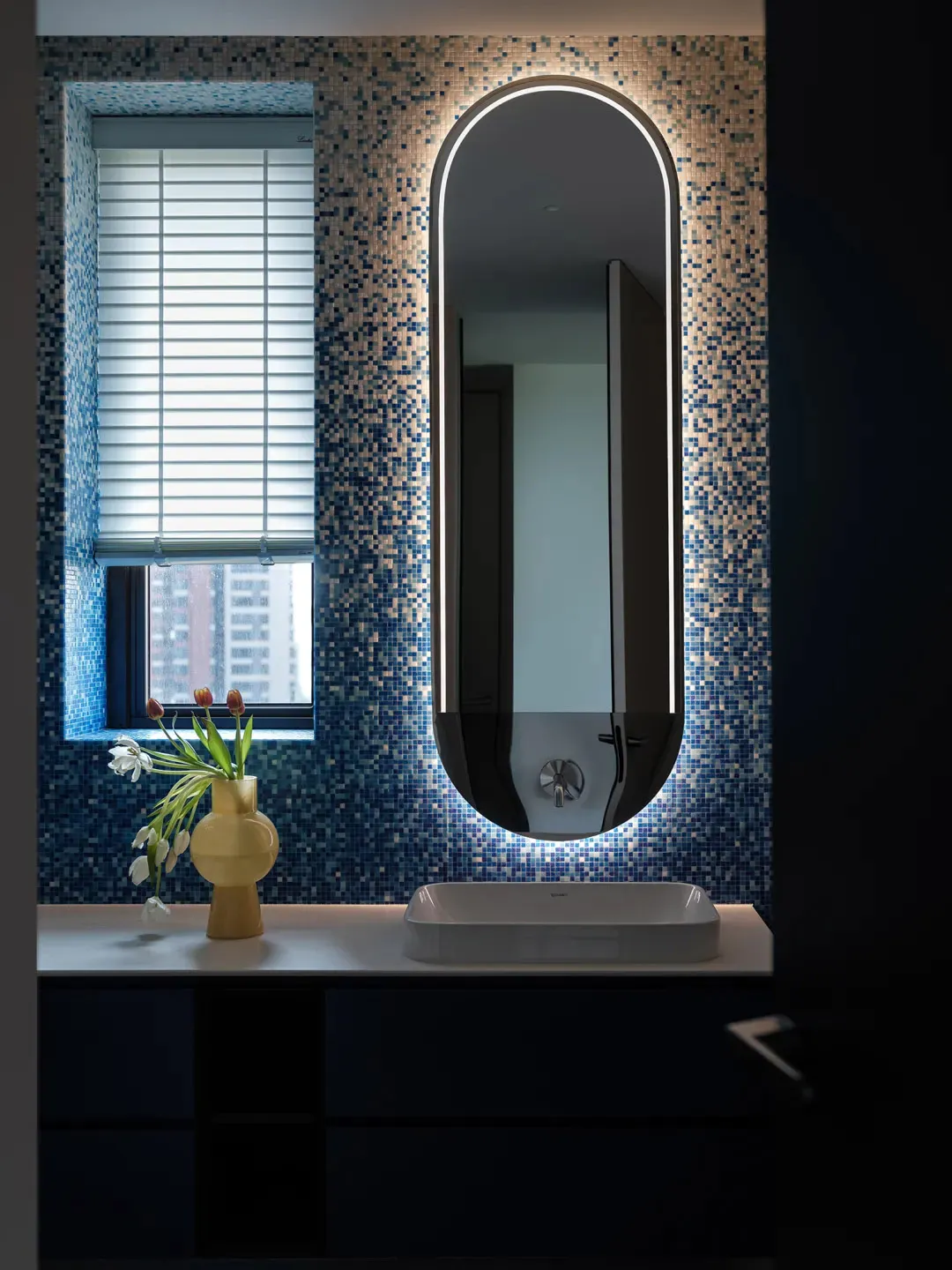 Bathroom with a white sink, mirror, and window, featuring vases with flowers and a clean, organized atmosphere