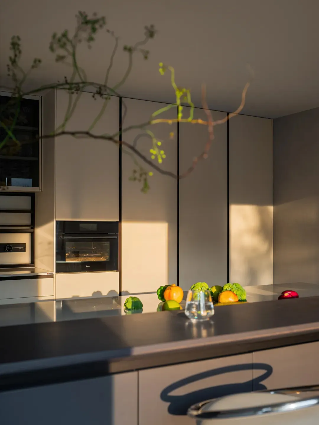 A clean and organized kitchen with white cabinets, a counter with apples and an orange, and a wine glass, set against a neutral background