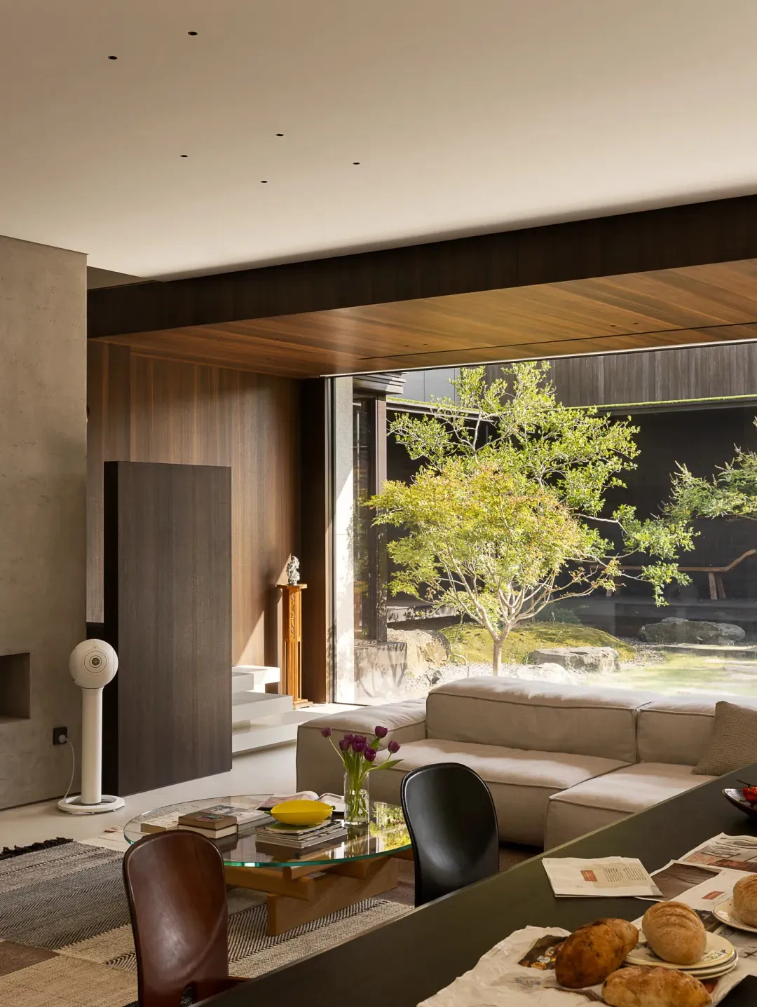 Modern living room with a couch, chair, dining table, and natural light from a large window, surrounded by books and clean lines