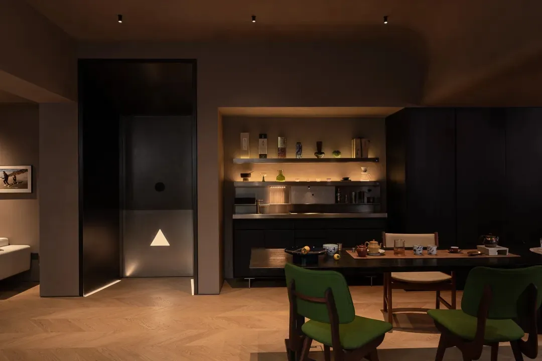 A modern kitchen dining space with a wooden floor, black cabinets, and warm lighting, featuring a dining table and chairs, cups, bowls, and vases