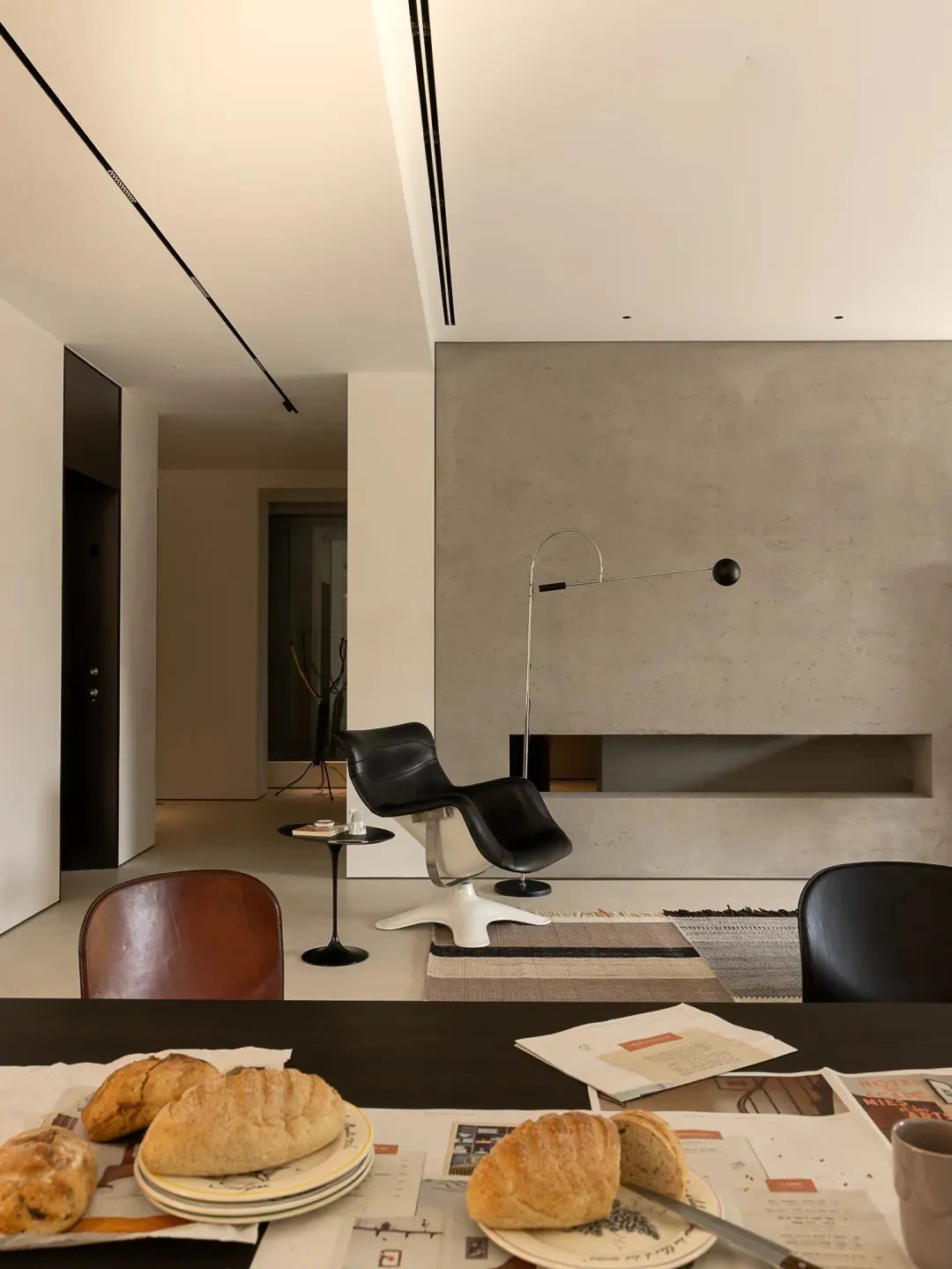 A modern living space with a dining table, chairs, and a couch, surrounded by concrete walls, with natural light and a cozy atmosphere